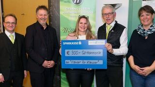 Fünf Personen stehen eng beieinander in einem Raum. Eine Frau hält einen blauen Scheck mit der Aufschrift Sparkasses und €900-GV-Harmonie. Sie lächelt in die Kamera. Hinter ihr steht ein Plakat, auf dem Gesangverein Harmonie Gotzis steht.
