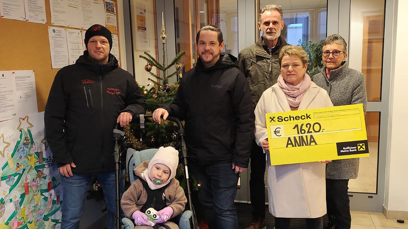 Eine Familie mit einem Baby im Kinderwagen steht vor einem geschmückten Weihnachtsbaum, wobei eine Frau ein gelbes Schild mit der Aufschrift 'Scheck € 1620 ANNA' hält.