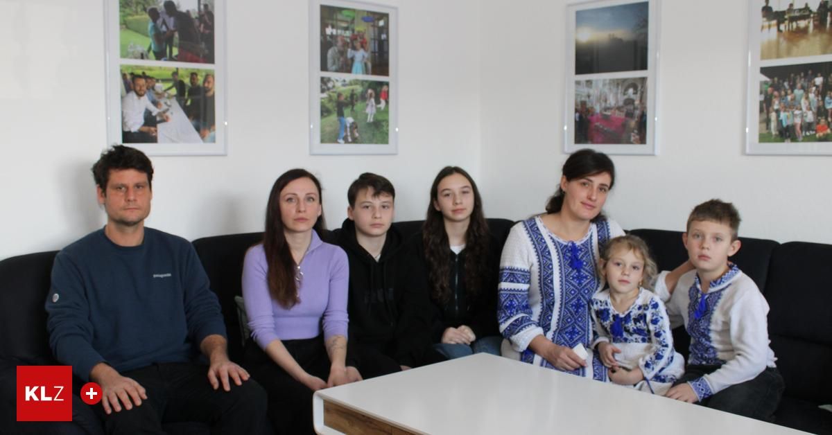 Eine Familie mit fünf Personen sitzt auf einer schwarzen Couch mit einem weißen Tisch davor. Die Frau rechts hält ein kleines Mädchen. Hinter ihnen hängen drei gerahmte Bilder an der Wand.