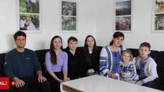 Eine Familie mit fünf Personen sitzt auf einer schwarzen Couch mit einem weißen Tisch davor. Die Frau rechts hält ein kleines Mädchen. Hinter ihnen hängen drei gerahmte Bilder an der Wand.