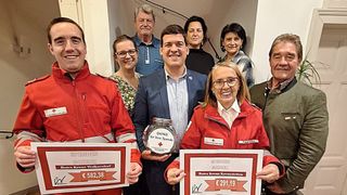Gruppe von Menschen posiert für ein Foto, zwei Frauen halten Spendenschecks und ein Mann hält einen Spendentopf, alle tragen rote Jacken, ein Mann in einem blauen Anzug, Innenraum mit Treppen im Hintergrund.