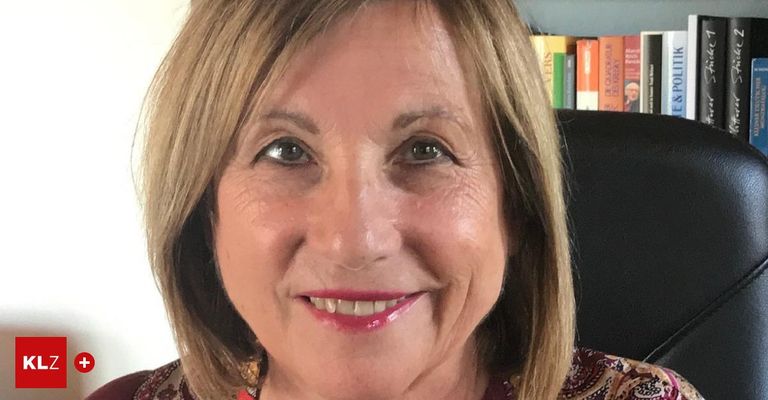 Eine lächelnde Frau mit kurzen blonden Haaren, in einem Blumentops, posiert vor einem Bücherregal mit verschiedenen Büchern.