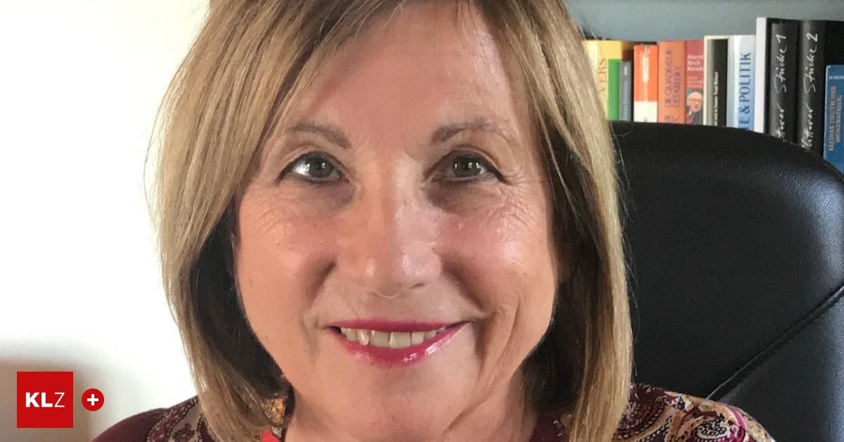 Eine lächelnde Frau mit kurzen blonden Haaren, in einem Blumentops, posiert vor einem Bücherregal mit verschiedenen Büchern.