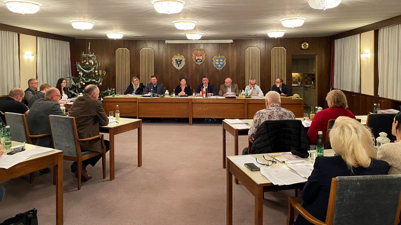 Eine Besprechung von Beamten in einem Konferenzraum mit einem Weihnachtsbaum in der Ecke. Mäntel sind auf dem Rücken der Stühle, und Mäntel sind auf dem Tisch.