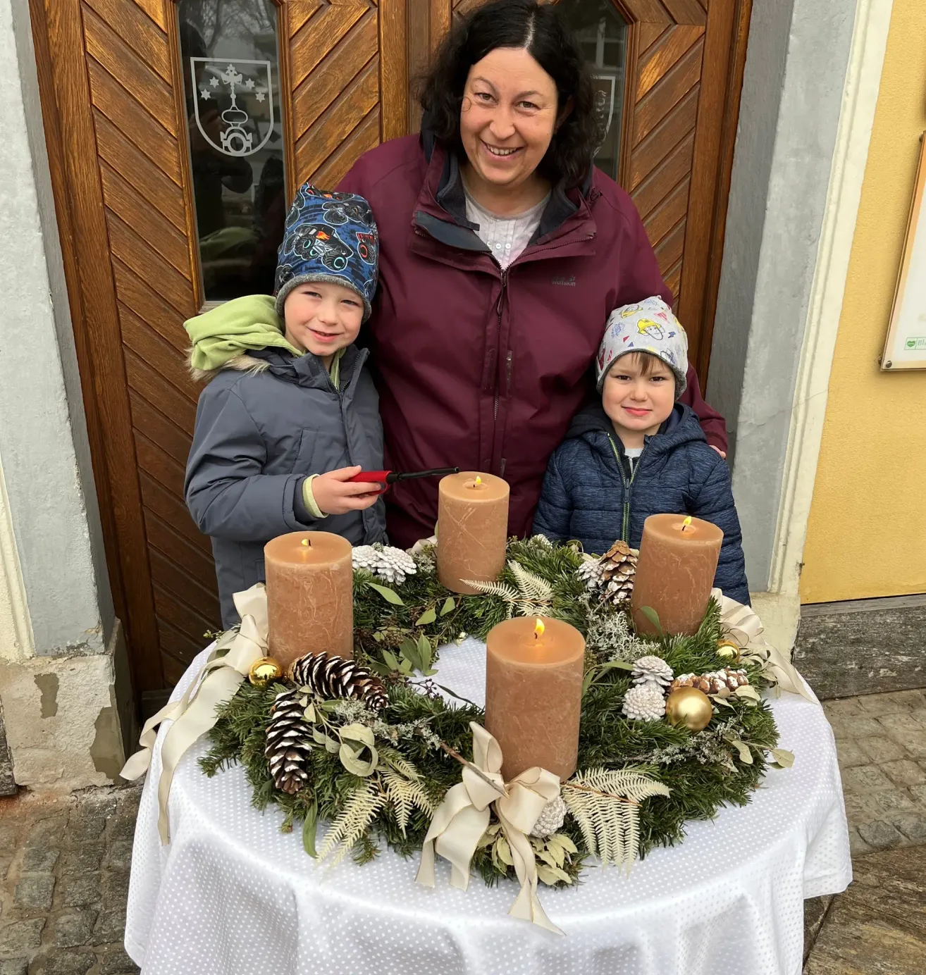 Eine lächelnde Frau steht mit zwei Kindern vor einem Kranz mit brennenden Kerzen. Der Kranz ist mit Tannenzapfen und einer weißen Schleife geschmückt. Die Kinder halten ein rotes Objekt.