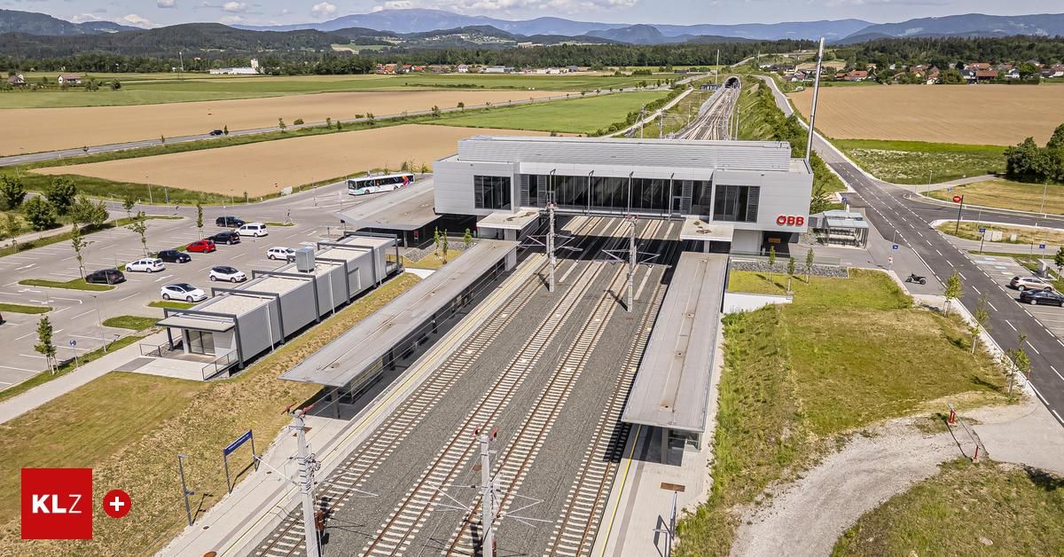 Luftaufnahme eines modernen Bahnhofs mit mehreren Gleisen, umgeben von einem Parkplatz und üppigen grünen Feldern. Berge in der Ferne.