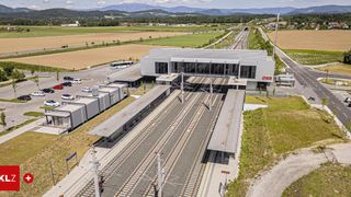 Luftaufnahme eines modernen Bahnhofs mit mehreren Gleisen, umgeben von einem Parkplatz und üppigen grünen Feldern. Berge in der Ferne.