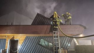 Feuerwehrleute stehen auf einer Leiter und auf dem Dach eines Gebäudes und bekämpfen einen Brand. Sie sind mit Helmen und Sauerstoffflaschen ausgestattet.