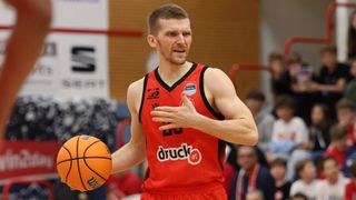 Ein Basketballspieler in einem roten Trikot mit dem Wort Druck drauf dribbelt den Ball. Er befindet sich auf einem Hallenplatz und ein Publikum beobachtet.