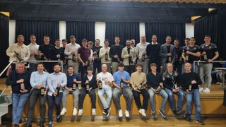 Eine Gruppe von Männern in unterschiedlicher Kleidung hält Flaschen und sitzt oder steht auf einer Holzbank mit Vorhanghintergrund.