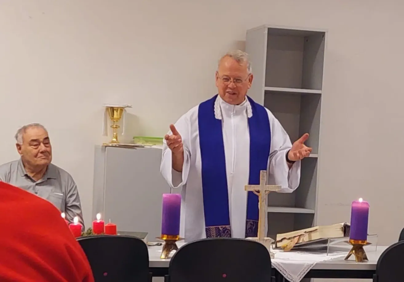 Ein Mann in einer weißen und blauen Robe steht vor einem Tisch mit Kerzen und einem Kreuz und spricht zu einem Publikum.