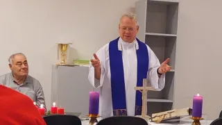 Ein Mann in einer weißen und blauen Robe steht vor einem Tisch mit Kerzen und einem Kreuz und spricht zu einem Publikum.