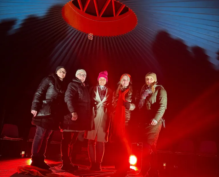Fünf Personen stehen in einem rot beleuchteten Raum. Sie tragen Winterkleidung. Eine trägt Hut und Schal, eine andere einen roten Hut. Der Raum hat eine rote Decke.