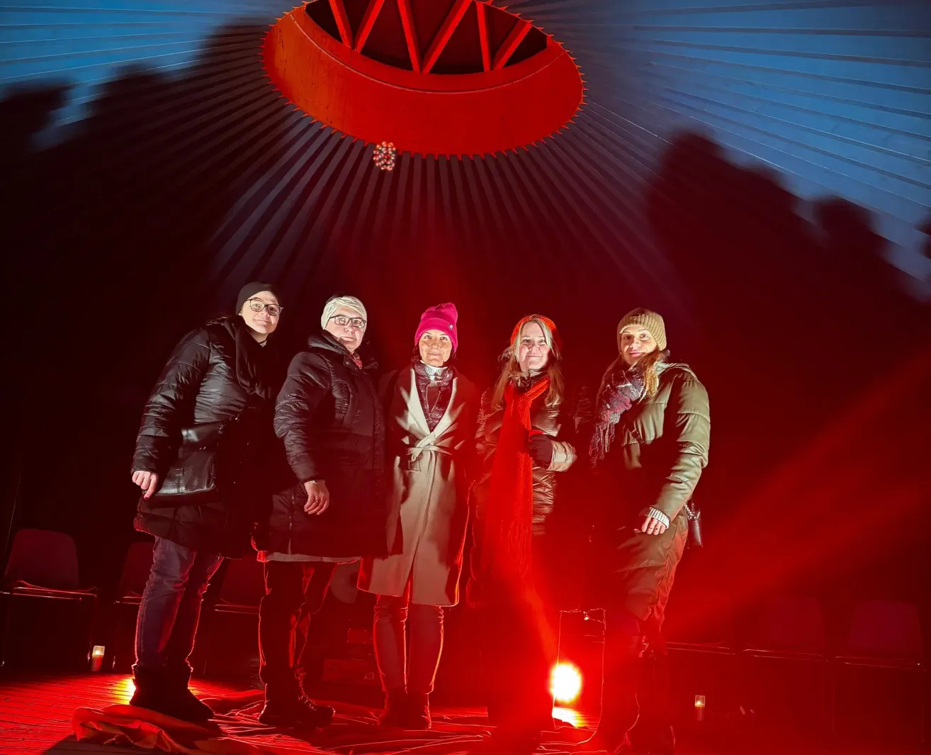 Fünf Personen stehen in einem rot beleuchteten Raum. Sie tragen Winterkleidung. Eine trägt Hut und Schal, eine andere einen roten Hut. Der Raum hat eine rote Decke.