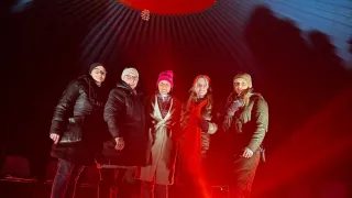 Fünf Personen stehen in einem rot beleuchteten Raum. Sie tragen Winterkleidung. Eine trägt Hut und Schal, eine andere einen roten Hut. Der Raum hat eine rote Decke.