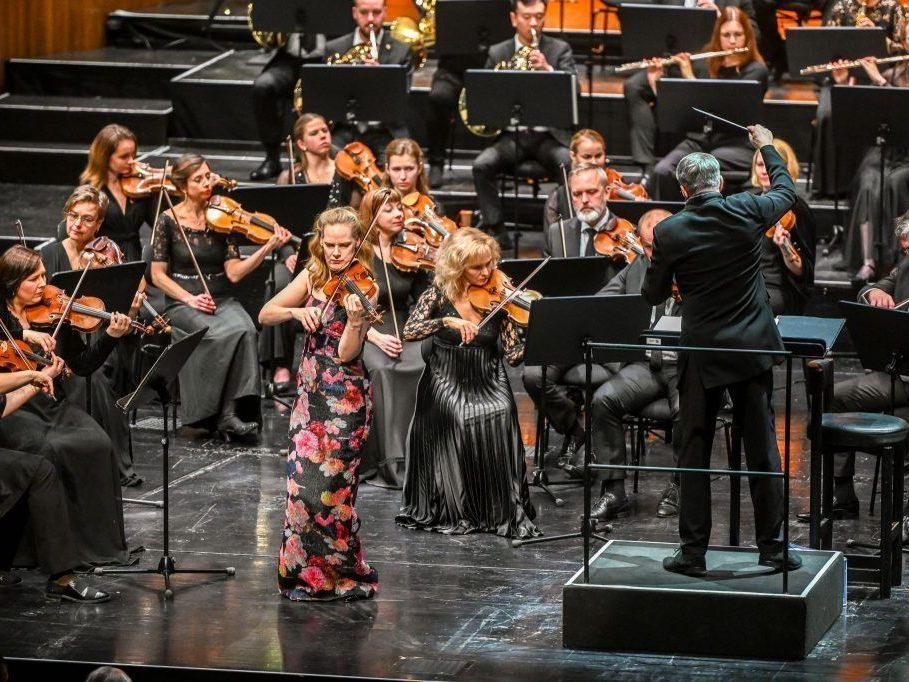 Ein Orchester auf der Bühne mit einem Dirigenten, Geigerinnen vorne und Musikern, die verschiedene Instrumente im Hintergrund spielen.