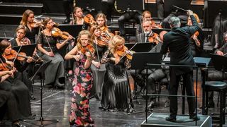 Ein Orchester auf der Bühne mit einem Dirigenten, Geigerinnen vorne und Musikern, die verschiedene Instrumente im Hintergrund spielen.