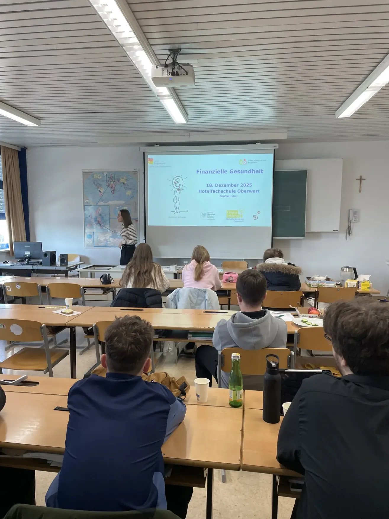 Ein Klassenzimmer mit Schülern in Reihen, die vor einer Projektionsleinwand sitzen, auf der eine Präsentation mit dem Titel 'Finanzielle Gesundheit' läuft. Eine Lehrerin steht in der Nähe der Leinwand, während die Schüler verschiedene Gegenstände auf ihren Tischen haben, darunter eine Tasse und eine Flasche.
