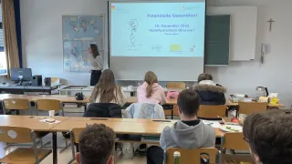 Ein Klassenzimmer mit Schülern in Reihen, die vor einer Projektionsleinwand sitzen, auf der eine Präsentation mit dem Titel 'Finanzielle Gesundheit' läuft. Eine Lehrerin steht in der Nähe der Leinwand, während die Schüler verschiedene Gegenstände auf ihren Tischen haben, darunter eine Tasse und eine Flasche.