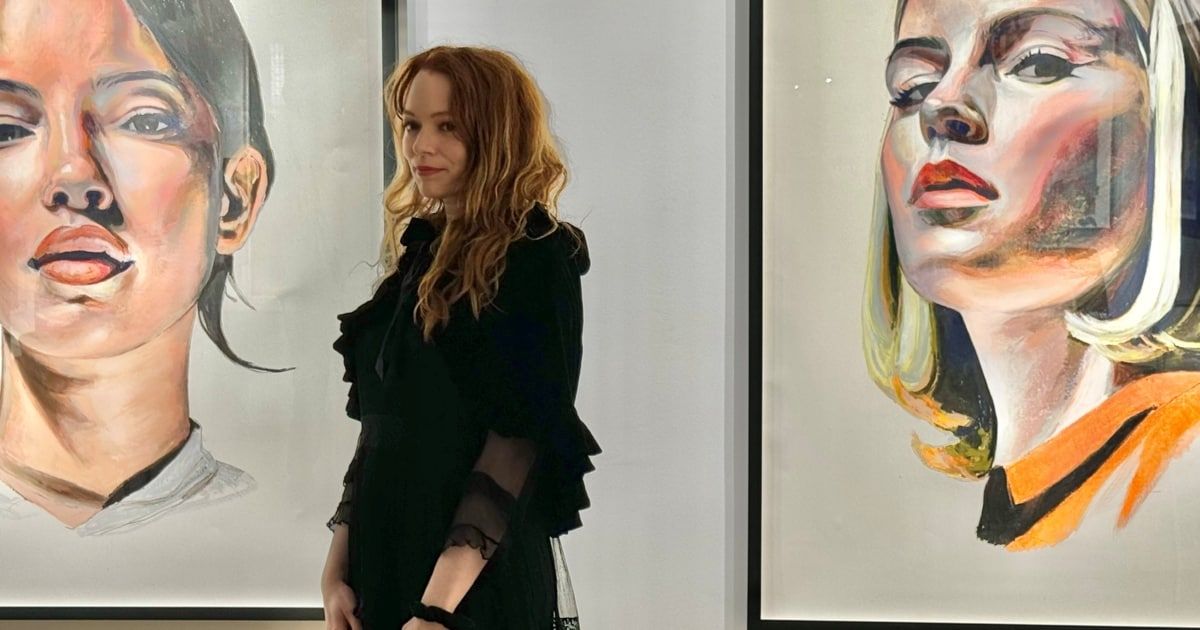 Eine Frau mit roten Haaren in einem schwarzen Kleid steht neben zwei großen Gemälden an einer weißen Wand.