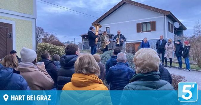 Eine Gruppe von Musikern spielt Blasinstrumente vor einem Haus. Das Publikum, bestehend aus Männern und Frauen, steht um sie herum. Die Umgebung ist draußen und der Himmel ist bewölkt.