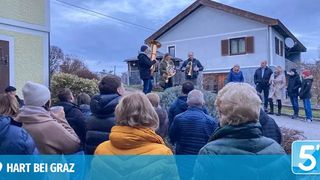 Eine Gruppe von Musikern spielt Blasinstrumente vor einem Haus. Das Publikum, bestehend aus Männern und Frauen, steht um sie herum. Die Umgebung ist draußen und der Himmel ist bewölkt.