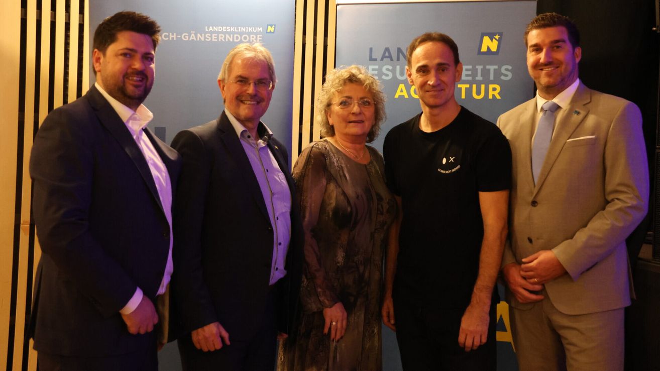 Eine Gruppe von vier Personen, schick gekleidet, steht vor einem blauen Hintergrund mit Text und Logo. Sie scheinen zu lächeln und posieren für ein Foto.