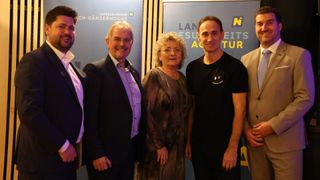 Eine Gruppe von vier Personen, schick gekleidet, steht vor einem blauen Hintergrund mit Text und Logo. Sie scheinen zu lächeln und posieren für ein Foto.