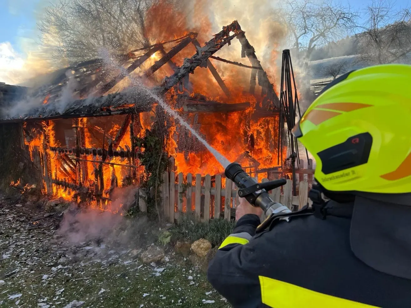 Ein Feuerwehrmann in voller Ausrüstung löscht ein brennendes Haus mit einem Schlauch, umgeben von Rauch und Flammen.