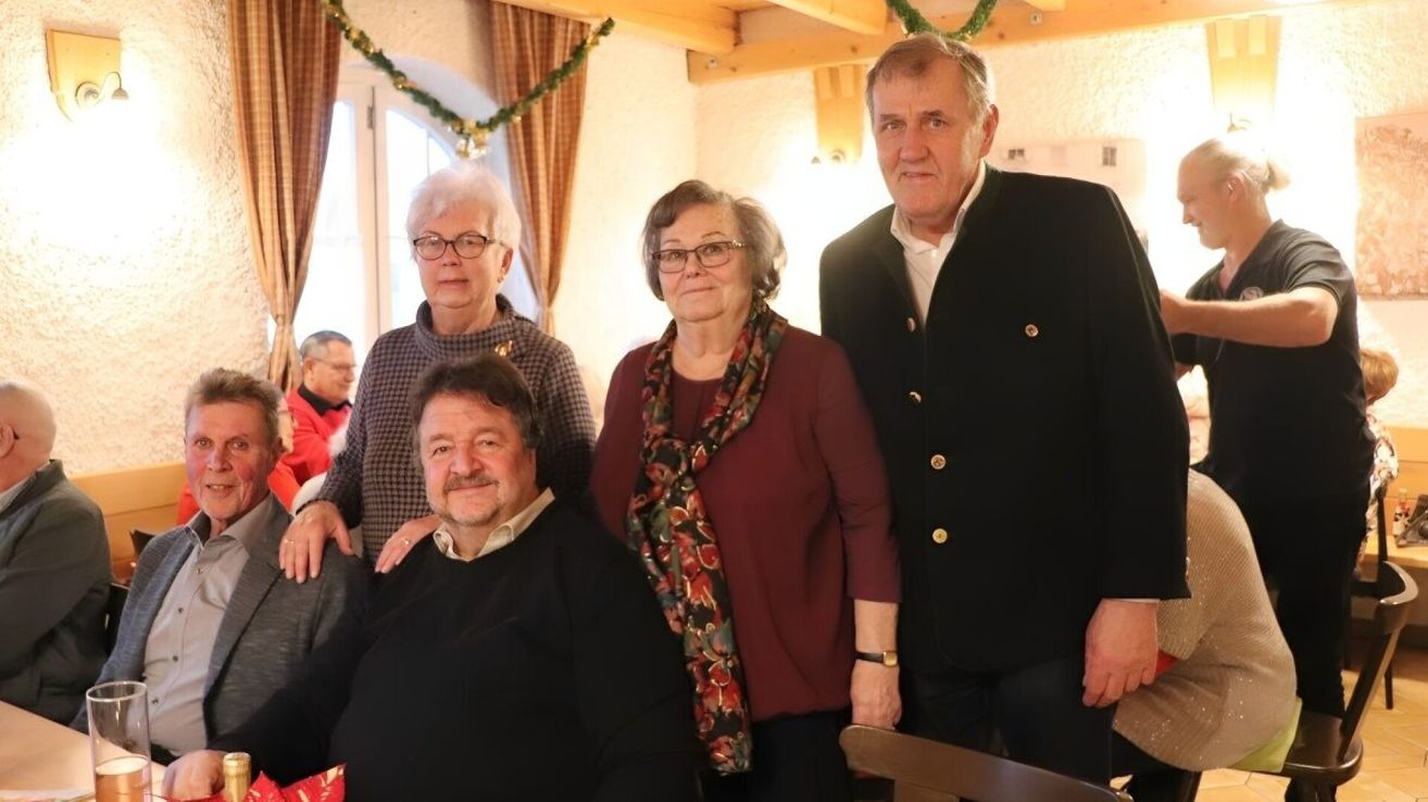 Vier Menschen stehen in einem Raum. Drei von ihnen posieren für ein Foto, während der vierte sitzt. Sie scheinen alle zu lächeln. Der Raum hat Holzbalken und Vorhänge.