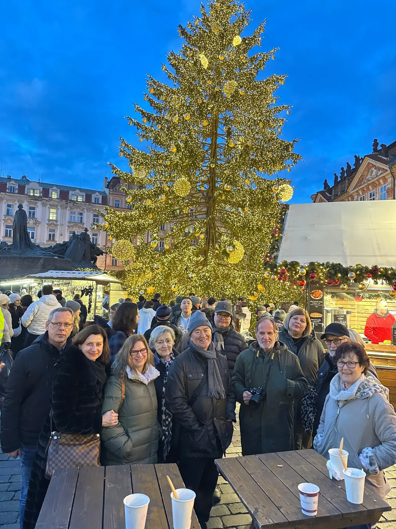 Eine Gruppe von Menschen posiert für ein Foto vor einem großen, geschmückten Weihnachtsbaum auf einem Stadtplatz. Hinter ihnen sind Gebäude mit Statuen und Straßenständen zu sehen.
