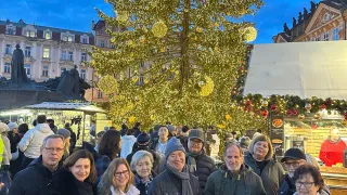 Eine Gruppe von Menschen posiert für ein Foto vor einem großen, geschmückten Weihnachtsbaum auf einem Stadtplatz. Hinter ihnen sind Gebäude mit Statuen und Straßenständen zu sehen.