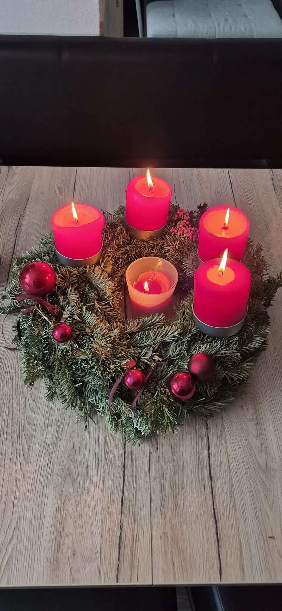 Ein Kranz mit vier rosa Kerzen auf einer Holzoberfläche, geschmückt mit roten Ornamenten und Grün. Die Kerzen sind angezündet, und der Kranz ist für festliche Feiern bestimmt.