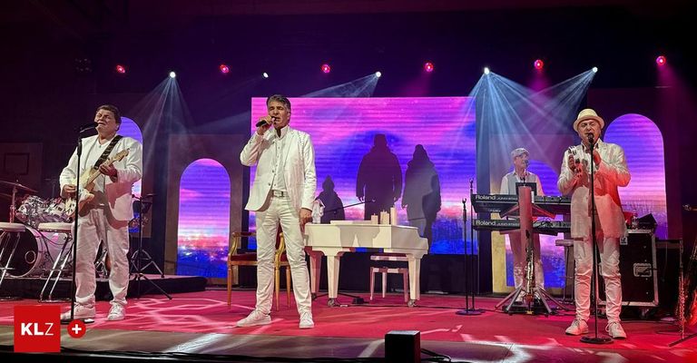 Ein Mann in einem weißen Anzug singt auf der Bühne vor einem Flügel. Dahinter befindet sich eine Leinwand mit Silhouetten. Zwei weitere Musiker spielen auf der Seite der Bühne Keyboards.