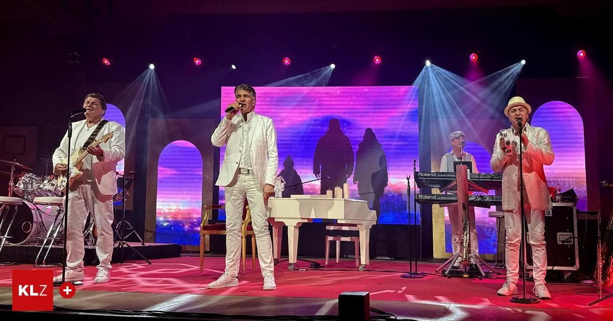 Ein Mann in einem weißen Anzug singt auf der Bühne vor einem Flügel. Dahinter befindet sich eine Leinwand mit Silhouetten. Zwei weitere Musiker spielen auf der Seite der Bühne Keyboards.