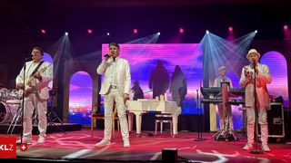 Ein Mann in einem weißen Anzug singt auf der Bühne vor einem Flügel. Dahinter befindet sich eine Leinwand mit Silhouetten. Zwei weitere Musiker spielen auf der Seite der Bühne Keyboards.