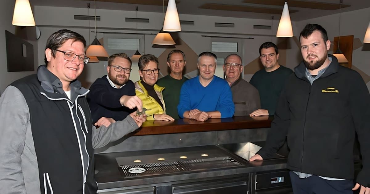 Eine Gruppe von Männern posiert für ein Foto an einer Küchentheke, einer hält ein kleines Dinosauriermodell, alle lächeln.