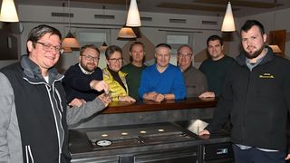 Eine Gruppe von Männern posiert für ein Foto an einer Küchentheke, einer hält ein kleines Dinosauriermodell, alle lächeln.