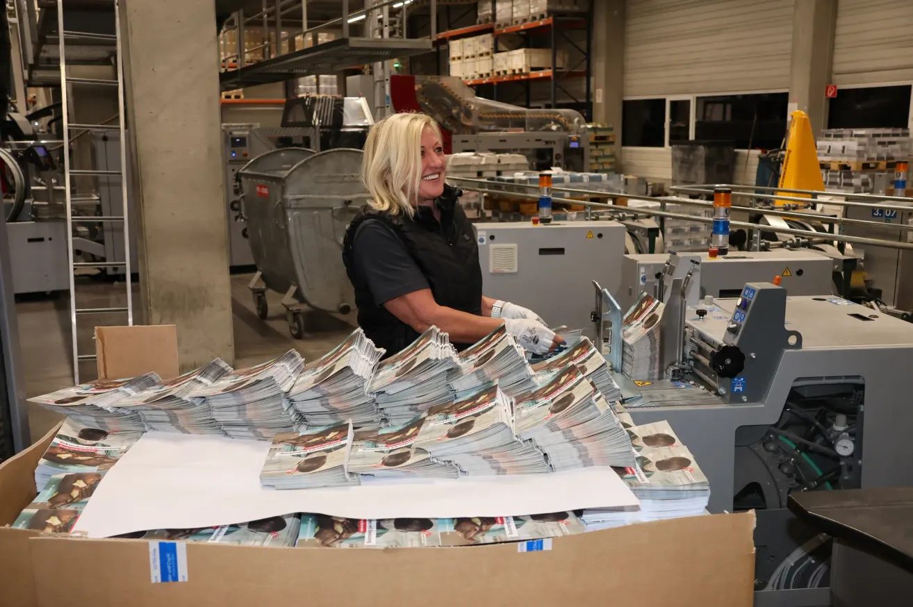 Eine Frau in einer Fabrikumgebung mit Handschuhen und einem schwarzen Shirt lächelt und arbeitet an einer Maschine mit einem großen Stapel Zeitschriften.