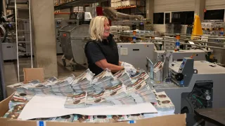 Eine Frau in einer Fabrikumgebung mit Handschuhen und einem schwarzen Shirt lächelt und arbeitet an einer Maschine mit einem großen Stapel Zeitschriften.