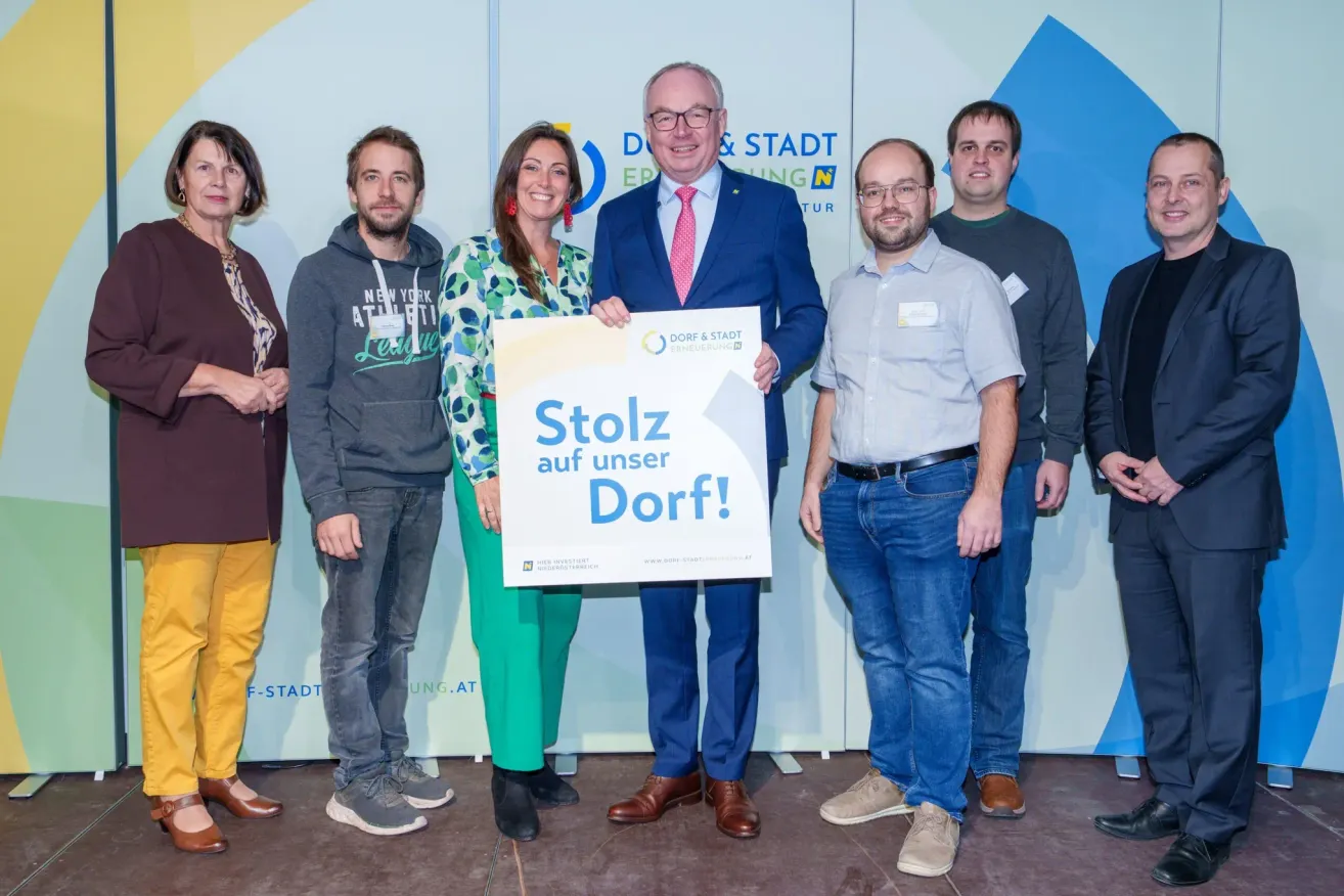 Eine Gruppe von Menschen posiert für ein Foto mit einem Schild, auf dem Stolz auf unser Dorf! steht. Der Mann in der Mitte hält das Schild und alle tragen Freizeitkleidung. Hinter ihnen ist eine Wand mit einem blau-weißen Design.
