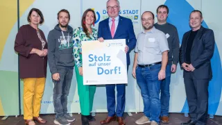 Eine Gruppe von Menschen posiert für ein Foto mit einem Schild, auf dem Stolz auf unser Dorf! steht. Der Mann in der Mitte hält das Schild und alle tragen Freizeitkleidung. Hinter ihnen ist eine Wand mit einem blau-weißen Design.