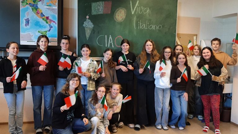 Eine Gruppe junger Frauen und Mädchen mit italienischen Flaggen steht vor einer Tafel mit italienischen Phrasen und einer Zeichnung eines Eisbechers.