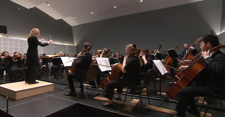 Ein Orchester spielt auf einer Bühne, mit Musikern, die verschiedene Instrumente wie Cellos und Geigen spielen. Der Dirigent steht vorne und leitet das Ensemble.