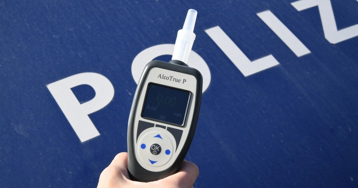 Eine Hand hält ein AlcoTrue P-Gerät vor einem blauen Hintergrund mit weißen Buchstaben, die 'POLI' buchstabieren. Das Gerät zeigt '0,00' auf dem Bildschirm an.