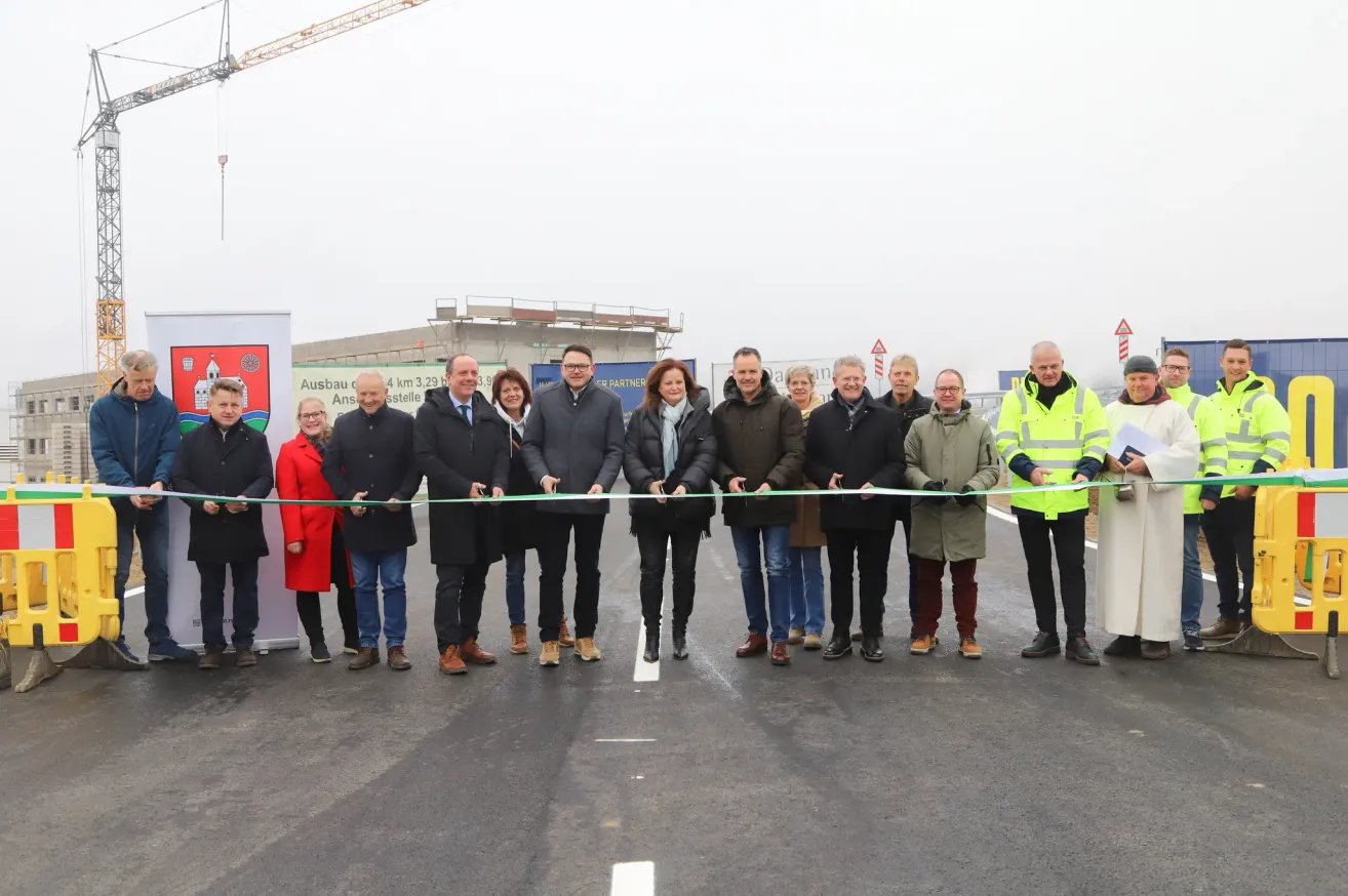 Eine Gruppe von Menschen in Mänteln steht draußen und hält ein grünes Band, vor einer Baustelle.