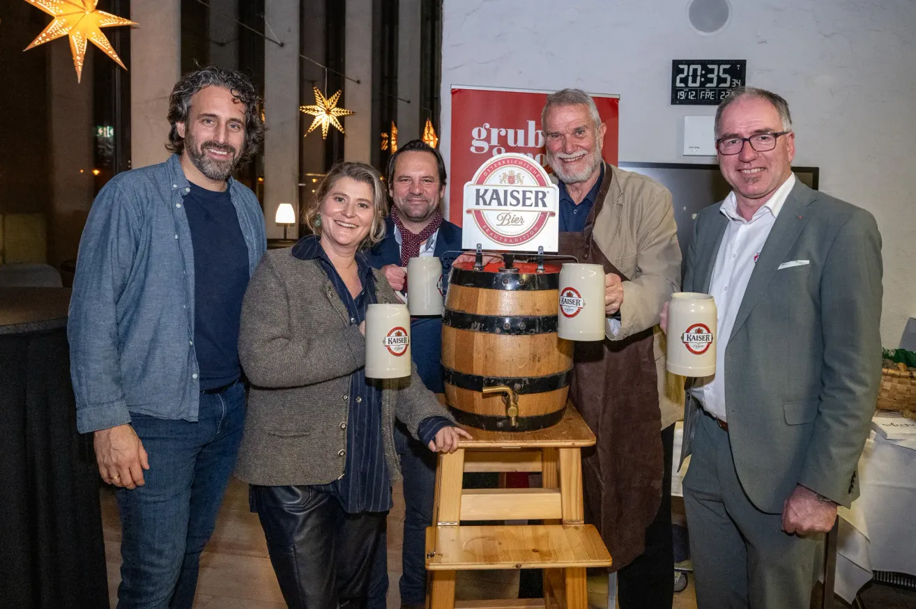Fünf Erwachsene, zwei Männer und drei Frauen, posieren mit Bierkrügen um einen Holzfass, das mit Kaiser Bier beschriftet ist. Sie stehen in einem Raum mit einer digitalen Uhr und festlichen Dekorationen.