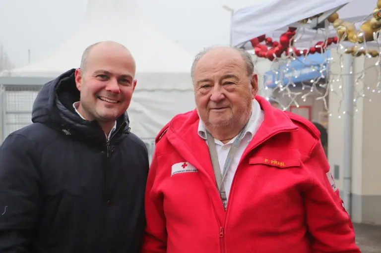 Zwei Männer stehen lächelnd für ein Foto. Der Mann auf der linken Seite trägt eine schwarze Jacke, während der Mann auf der rechten Seite eine rote Jacke mit einem roten Kreuzemblem trägt.