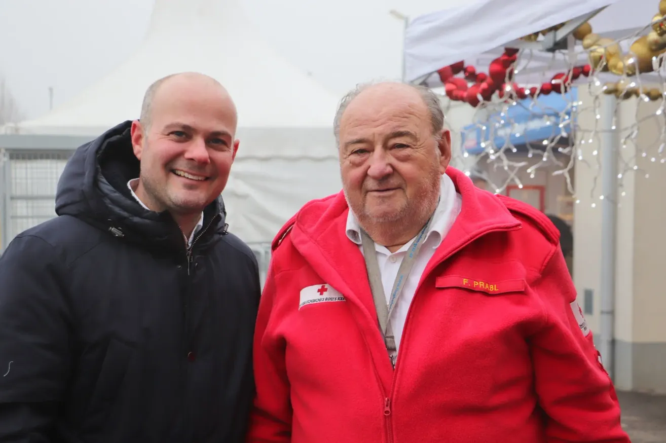 Zwei Männer stehen lächelnd für ein Foto. Der Mann auf der linken Seite trägt eine schwarze Jacke, während der Mann auf der rechten Seite eine rote Jacke mit einem roten Kreuzemblem trägt.