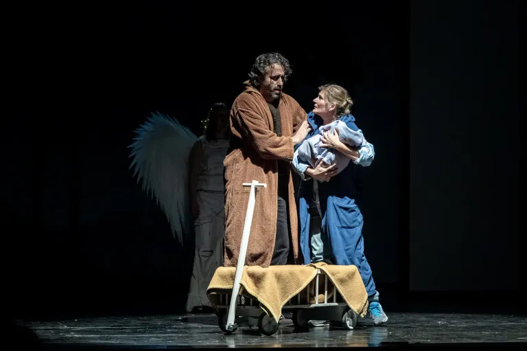 Zwei Menschen stehen auf der Bühne und halten ein Baby. Eine Person mit Flügeln steht hinter ihnen. Die Bühne ist dunkel.